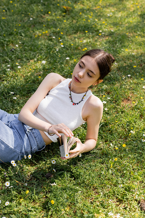 High angle view of young brunette woman in top and jeans holding book while lying on lawn with daisies in park