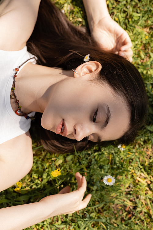Top view of young brunette woman looking at flowers while lying on lawn in park