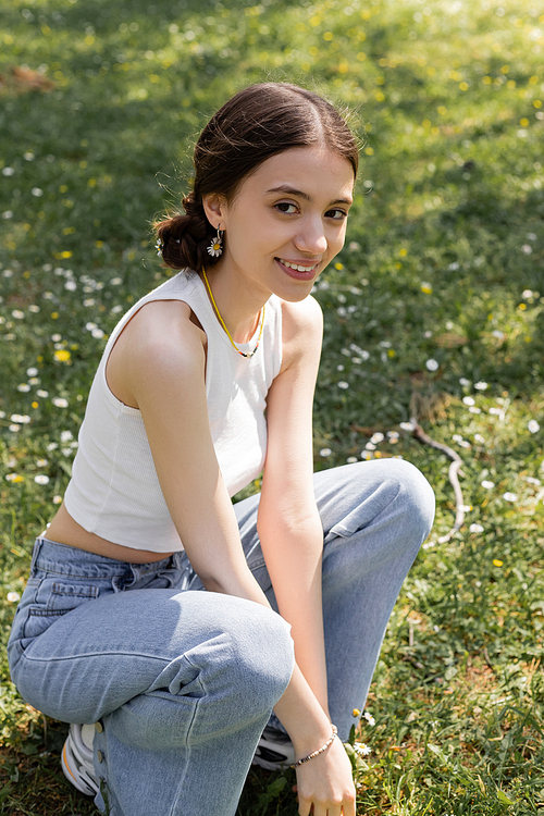 Carefree young woman in top and jeans looking at camera on meadow with flowers in park