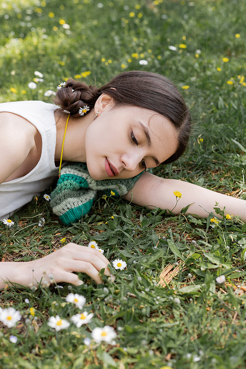 Portrait of young brunette woman looking at blurred daisies while lying on meadow in park