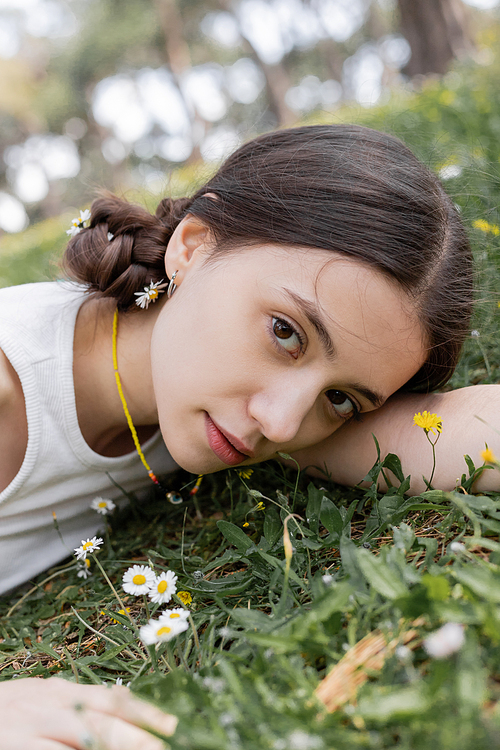 Portrait of young brunette woman looking at camera near daisies on lawn in park