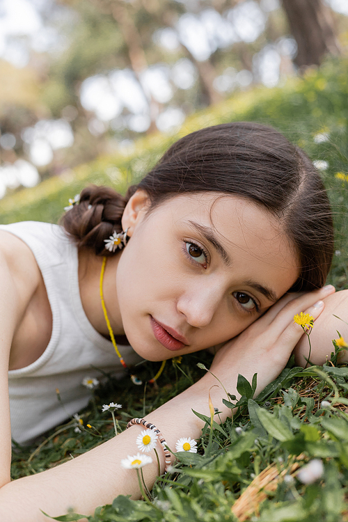 Portrait of pretty young woman looking at camera while lying on meadow with flowers in park