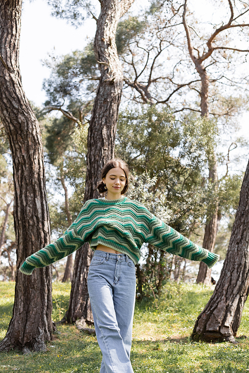 Young brunette woman in knitted sweater walking on lawn in summer park