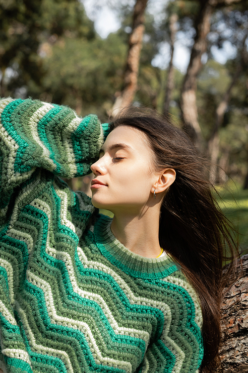 Young woman in knitted sweater touching hair near tree in park
