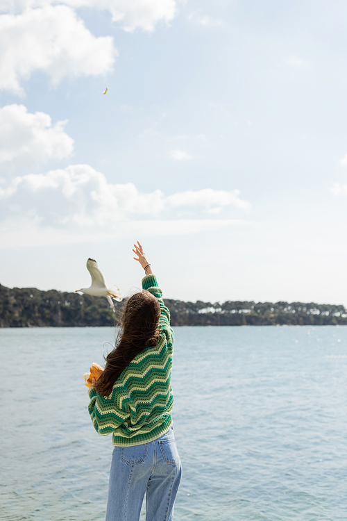 Back view of young woman holding bread near seagull flying over sea in Istanbul