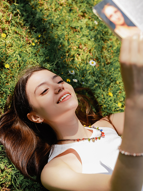 High angle view of smiling brunette woman reading book while lying on lawn with flowers