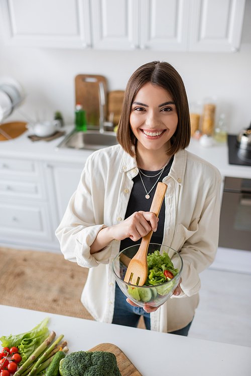 Positive young woman holding fresh salad and looking at camera in kitchen