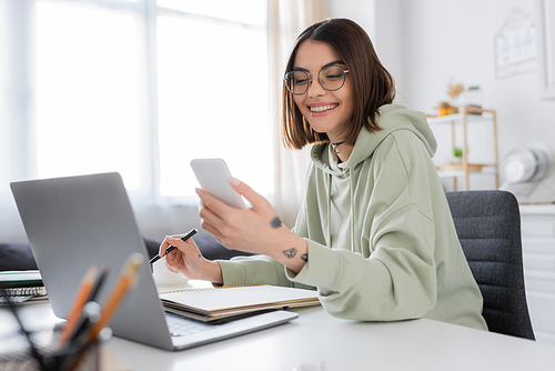 Cheerful young woman using smartphone and laptop near notebook at home