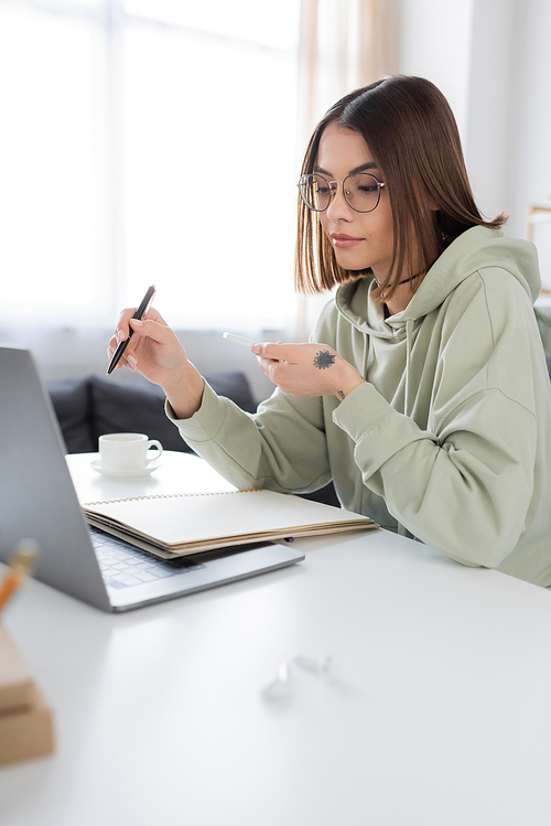 Brunette freelancer in eyeglasses holding smartphone near laptop and notebook on table at home