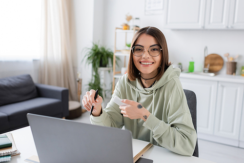 Smiling freelancer in eyeglasses holding smartphone and looking at camera near laptop at home
