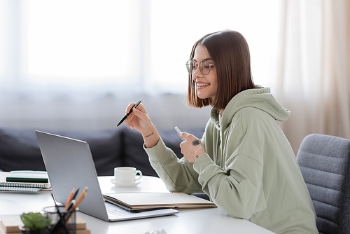 Positive freelancer in eyeglasses holding smartphone near laptop and coffee at home