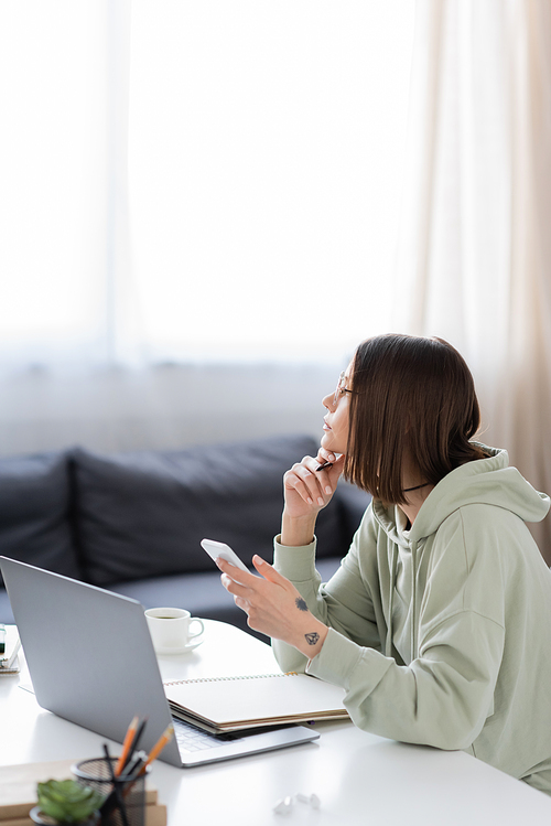 Side view of pensive freelancer holding cellphone near laptop and notebook at home