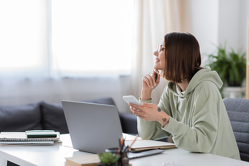 Side view of smiling tattooed freelancer using smartphone near laptop at home