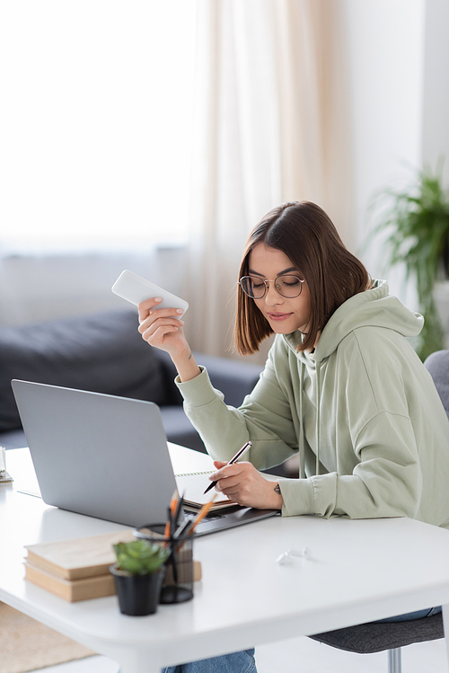 Young freelancer holding smartphone near laptop and writing on notebook at home