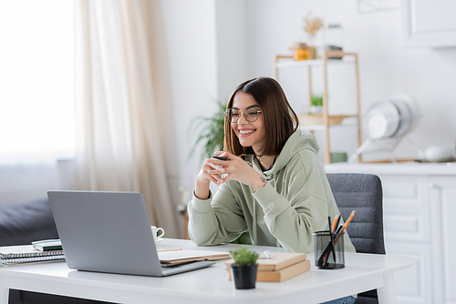 Smiling freelancer in eyeglasses holding pen near laptop and notebooks at home