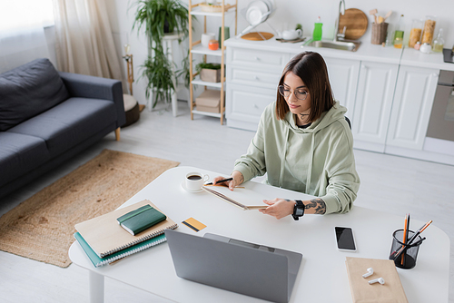 Tattooed freelancer holding notebook near devices and credit cart on table at home