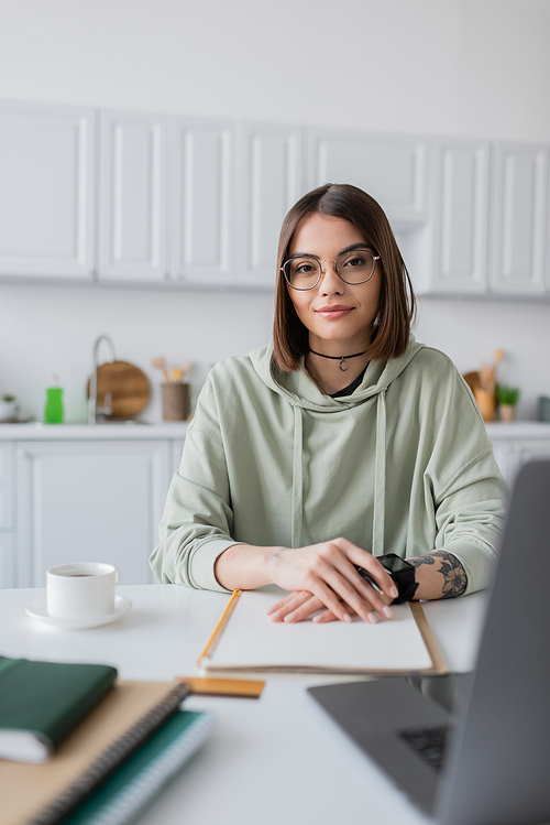 Young freelancer looking at camera near blurred laptop and coffee at home