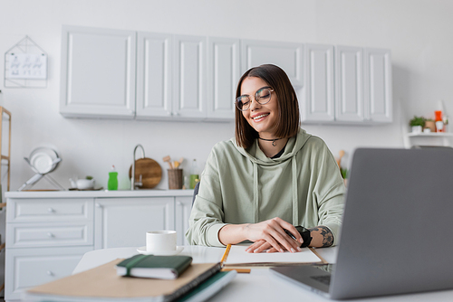Positive freelancer in eyeglasses looking at coffee cup near laptop and notebooks on table at home