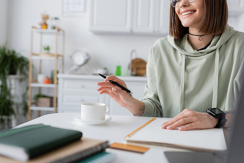 Cropped view of positive freelancer holding pen near coffee and notebooks at home