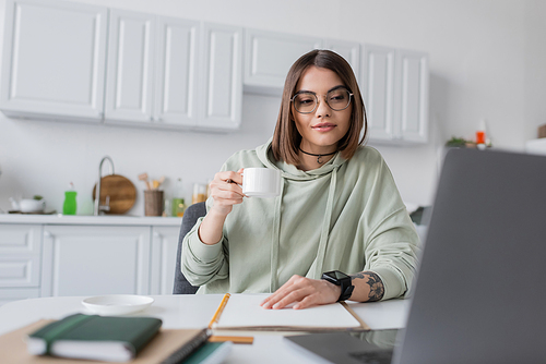 Tattooed freelancer in eyeglasses holding coffee near blurred laptop and notebooks at home