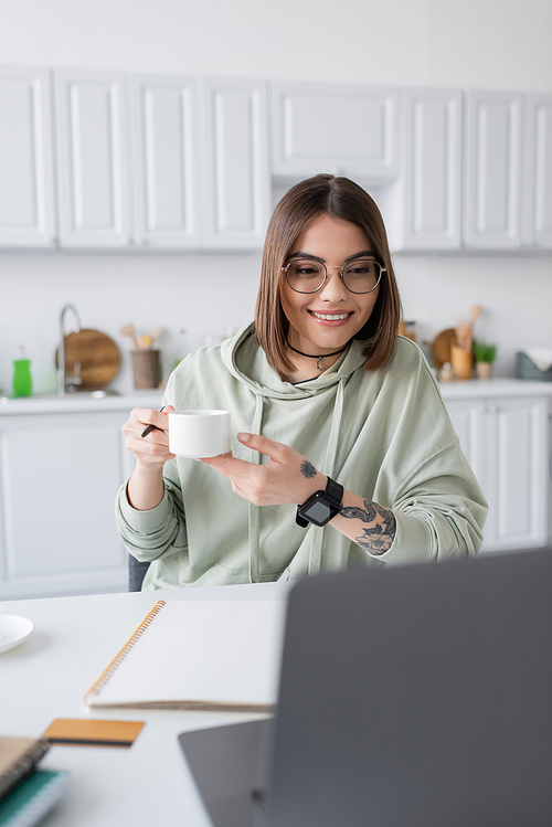 Cheerful freelancer holding coffee cup near blurred laptop and credit card at home