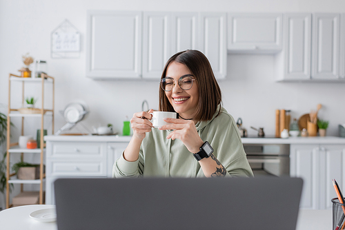 Positive freelancer in eyeglasses holding coffee near blurred laptop at home