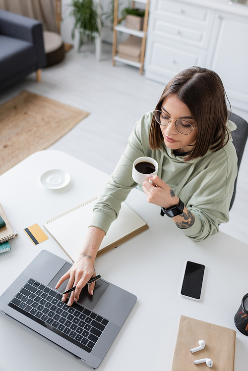 Overhead view of tattooed freelancer holding coffee and using laptop near notebooks and earphones at home