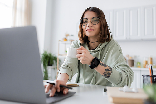 Tattooed freelancer in eyeglasses holding coffee cup and using blurred laptop at home