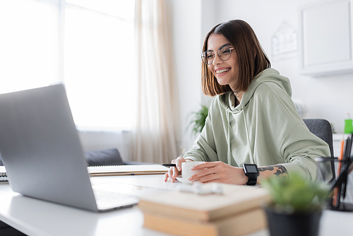 Cheerful freelancer in eyeglasses looking at laptop near coffee and books at home