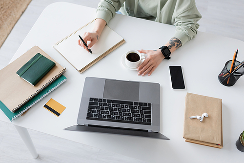 Top view of tattooed freelancer sitting near coffee and devices on table at home