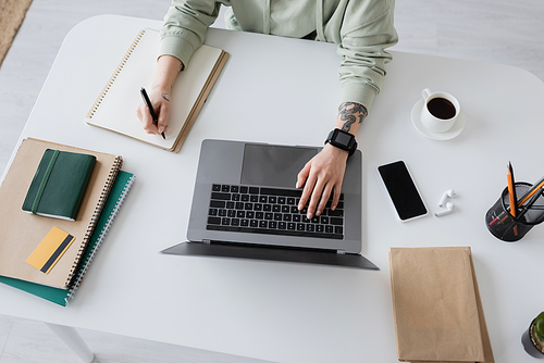 Cropped view of freelancer using laptop and writing on notebook near coffee and earphones on table at home