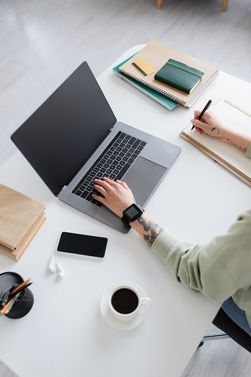 Top view of tattooed freelancer writing on notebook and using laptop near coffee and books on table