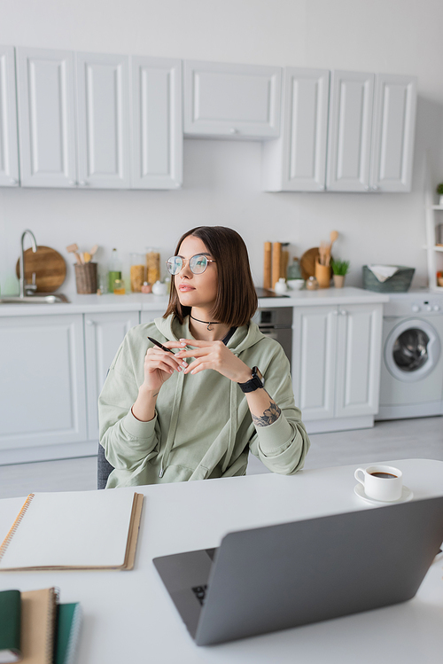 Young freelancer in eyeglasses holding pen near laptop and notebook on table at home