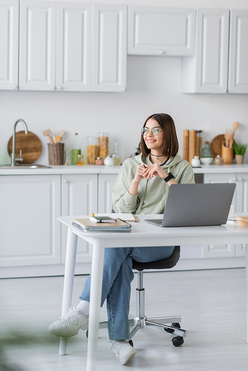 Smiling woman sitting near laptop and notebooks on table in kitchen