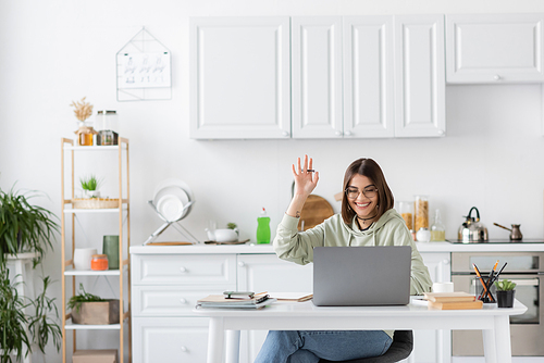 Positive freelancer having video call on laptop near coffee and notebooks on table at home