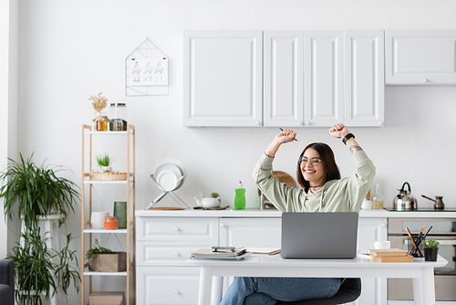 Excited freelancer showing yes gesture near laptop and notebooks on table in kitchen