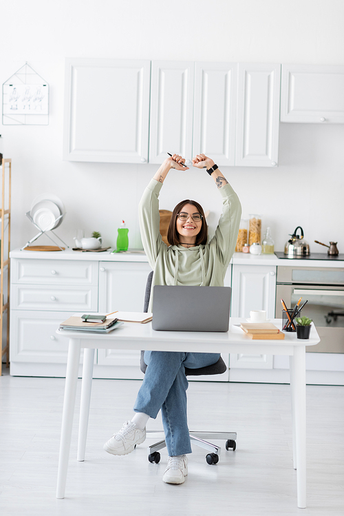 Excited freelancer in eyeglasses showing yes gesture near laptop and notebooks in kitchen