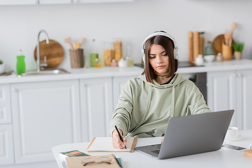 Young freelancer in headphones writing on notebook near gadgets and coffee on table at home