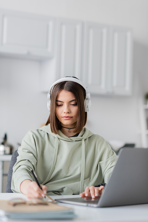 Brunette freelancer in headphones using laptop and blurred notebook at home