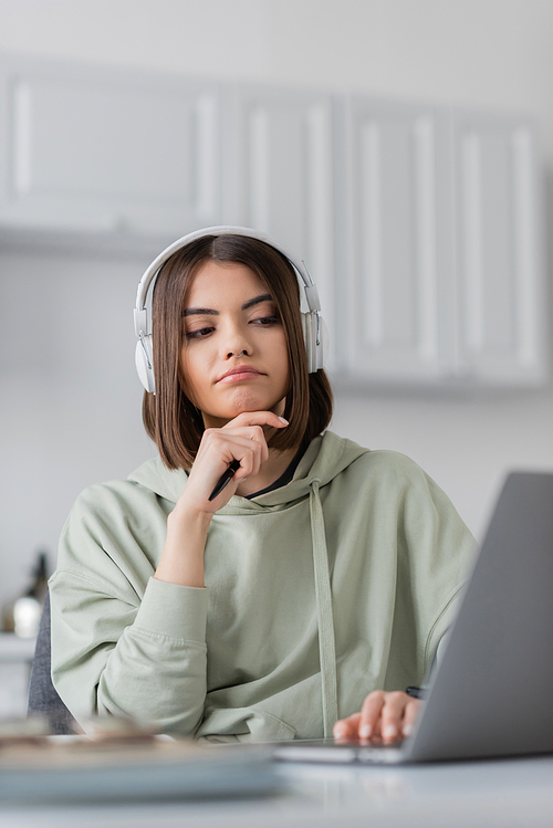 Pensive freelancer in headphones using blurred laptop at home