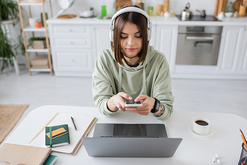 Brunette freelancer in headphones using smartphone near laptop and coffee at home
