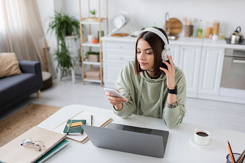 Young freelancer in headphones using smartphone near laptop and notebooks on table at home