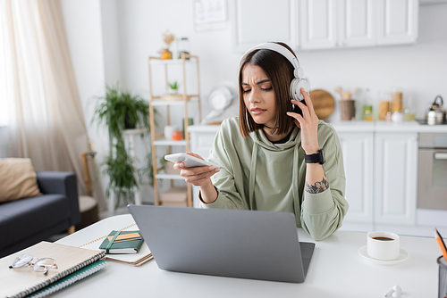 Displeased freelancer in headphones holding cellphone near laptop and coffee at home