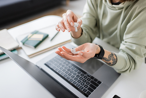 Cropped view of tattooed freelancer holding earphones near laptop with blank screen on table at home