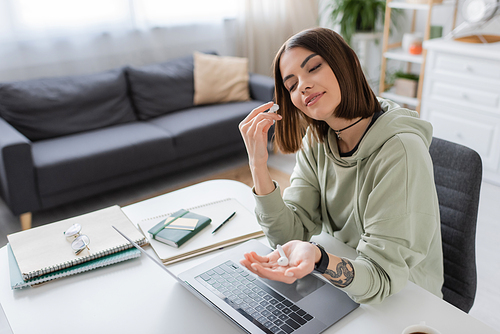 Smiling tattooed freelancer holding earphones near laptop and notebooks at home