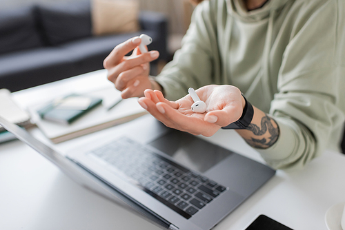 Cropped view of tattooed freelancer holding earphones near devices on table at home