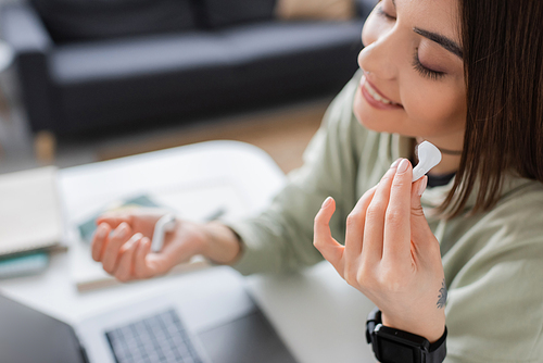 Cheerful freelancer holding earphone near blurred laptop at home