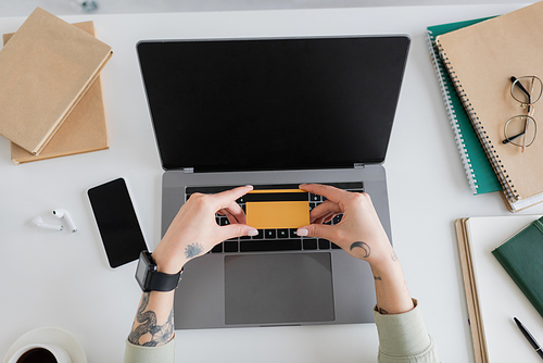 Top view of tattooed woman holding credit card near devices and notebooks on table at home