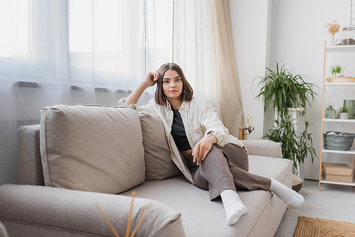 Brunette woman in casual clothes looking at camera while sitting on couch at home