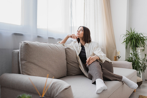 Young woman in casual clothes looking away while sitting on couch at home